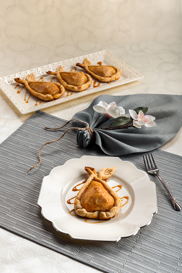 Gorgeous food photography of a dessert, Caramel Pie Pear. Beautifully plated on a silver trimmed ivory plate are the caramel pear pies. The plate is placed on a striped placemat in colors of blues, grays and ivory. A grayish silver fabric napkin is folded in a leaf shape, tied with twine and soft ivory flowers are tucked into the folds. A delicate lace trimmed tray with three Pear Pies also sets on a placemat. The set background is ivory with thin a delicately silver swirling pattern.