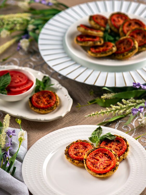oven roasted tomatoes on puff pastry rounds, a white plate plated with three Tomato Puffs and a sprig of basil, small oval white tray with small white bowl holding fresh sliced tomatoes, one tray with plated tomato puffs and accented with a sprig of basil.
