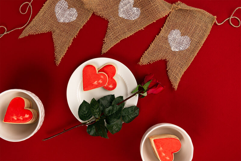 burlap heart banner, cookies and a single red rose setting on a red table cloth.